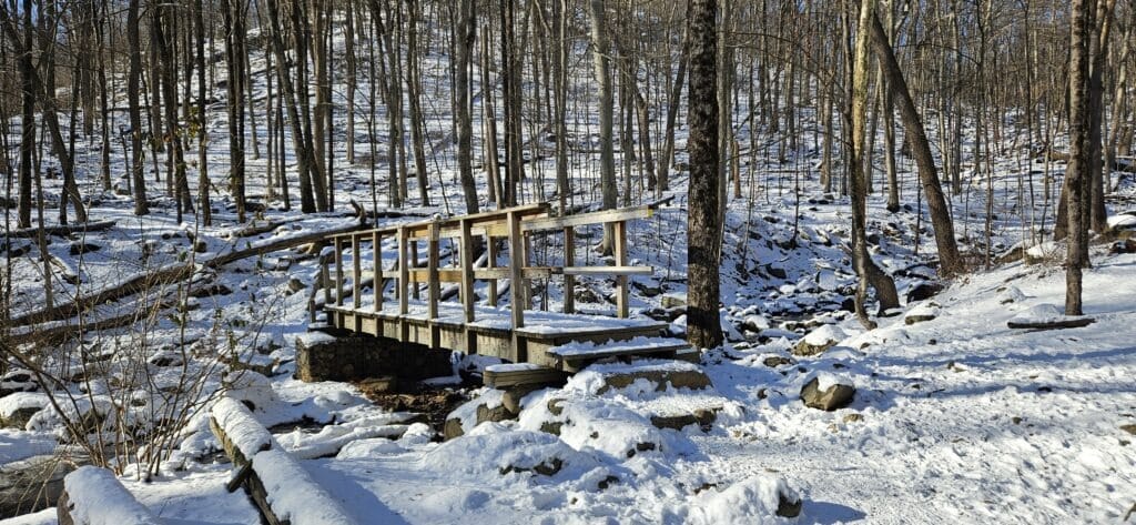 An image of the MacMillan Falls Trail in winter featuring a snow covered bridge